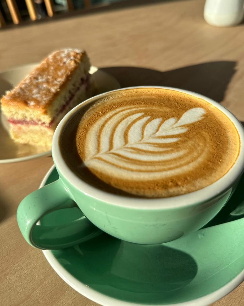 Flat white with rosetta latte art in a mint cup, a slice of sponge cake behind it.