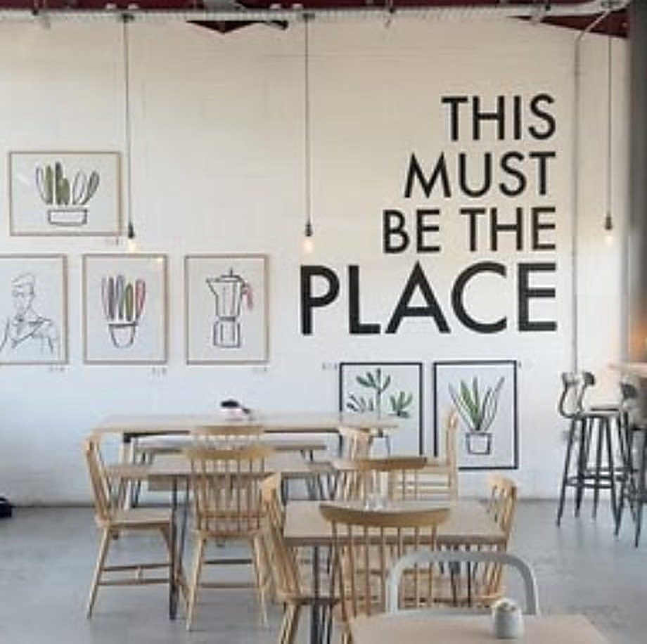 Light wooden tables and chairs under the mural, with framed plant and coffee-pot prints on the white wall.